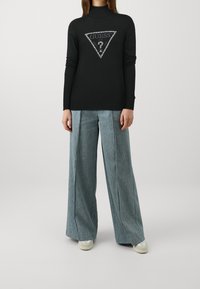 Maglione nero a collo alto con maniche lunghe e logo argentato a forma di triangolo. Abbinato a pantaloni azzurri a gamba larga e sneaker crema.