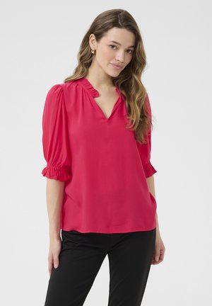 Blouse rose avec des manches bouffantes, un col volanté et un tissu doux. Associée à un pantalon noir, mettant en valeur une coupe décontractée.