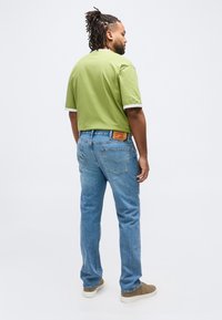 Άνδρας με dreadlocks στέκεται με την πλάτη, φορά ανοιχτό πράσινο μπλουζάκι με λευκή επένδυση, μπλε τζιν και μπεζ αθλητικά παπούτσια σε λευκό φόντο.