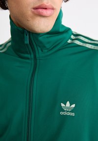 Zelena sportska jakna na patent, s visokim ovratnikom, koja ima tri bijele pruge na ramenima i mali bijeli logo na prsima.