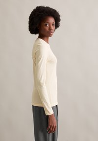 Blusa de manga longa, amarela clara, em corte justo, feita de um tecido suave. Combinada com calças cinzentas de riscas verticais, apresentando uma silhueta descontraída.