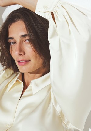 Blusa di satin crema con colletto classico, corpino aderente e maniche a sbuffo; texture morbida e leggero lucido. Dettagli con bottoni visibili.