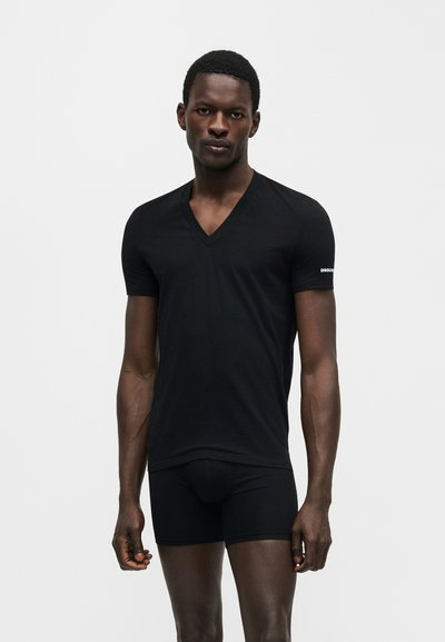 Άνδρας μοντέλο φοράει εφαρμοστή μαύρη μπλούζα με V λαιμόκοψη και μικρό λογότυπο στο μανίκι και μαύρο σορτς, στέκεται σε απλό φωτεινό φόντο.