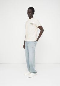 Μπεζ βαμβακερό t-shirt με πράσινο και κόκκινο λογότυπο από πάνω, συνδυασμένο με ανοιχτό μπλε φαρδιά τζιν και λευκά αθλητικά παπούτσια. Μινιμαλιστικό φόντο.