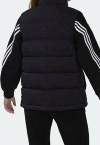 Svart pufferväst med hög krage, har vita tre-stripes accenter på ärmarna och en slät, matt textur.