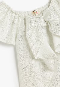 Top bianco scollato sulle spalle con tessuto testurizzato e lucido, colletto arricciato e un grande dettaglio a fiocco davanti.