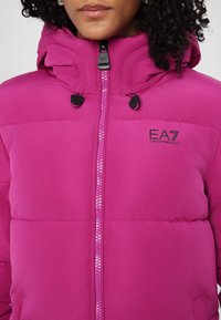 Rožinė pūkinė striukė su aukštu apykakle, priekiu užtrauktuku ir reguliuojama gaubtu. Kairėje krūtinėje yra EA7 Emporio Armani logotipas.
