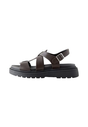 Smeđa kožna sandala s debelim crnim potplatom, ukriženim trakama i podesivom srebrnom kopčom na bijeloj pozadini.