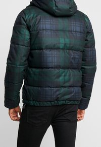 Puffjacka med ett rutmönster i grönt och mörkblått, vadderad design, fast huva och elastisk midja. Svarta byxor är påklädda under.