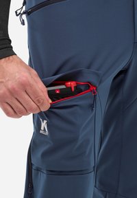 Pantalons millet bleu marine avec une poche zippée dotée d'une fermeture éclair rouge, montrant des outils à l'intérieur. Le matériau semble lisse avec une finition légèrement texturée.