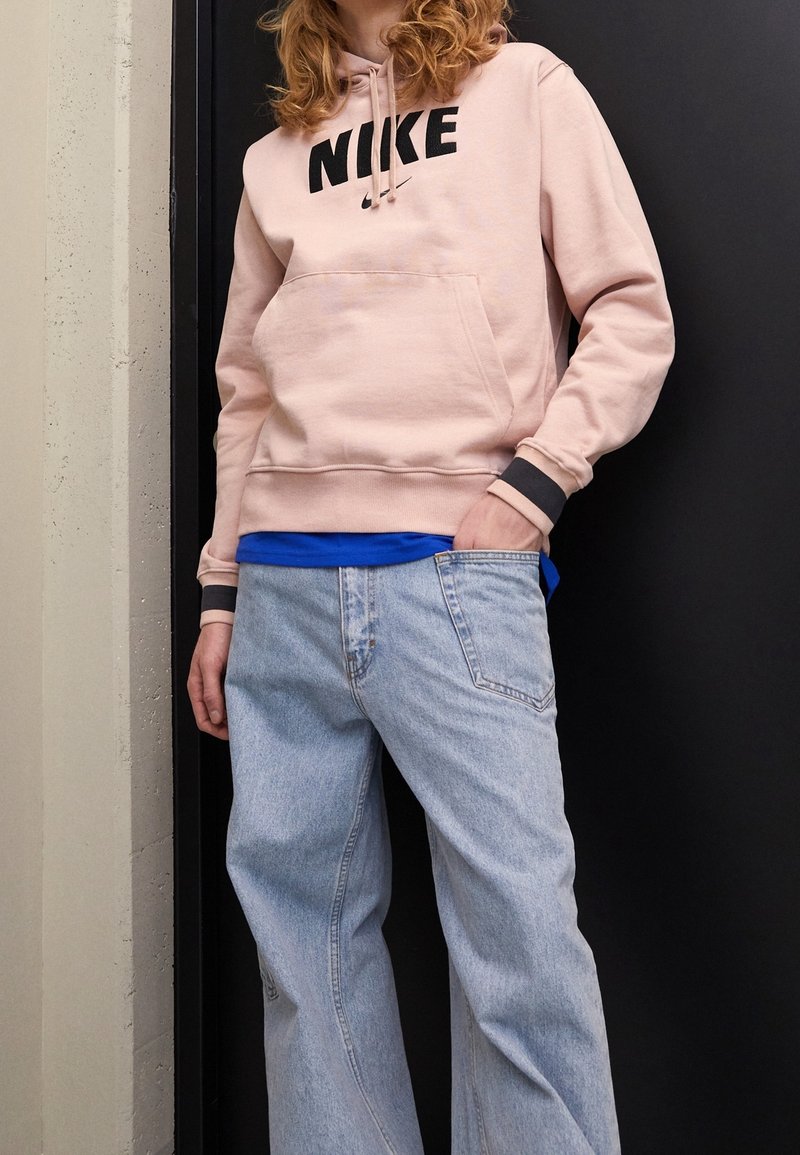 Sudadera rosa de Nike con logo negro, bolsillo canguro, puños grises, combinada con jeans baggy azul claro. Textura suave, diseño casual.
