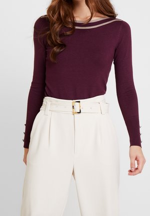 Femme portant un haut ajusté à manches longues de couleur violet foncé avec un détail beige au niveau de l'encolure, rentré dans un pantalon blanc taille haute avec une ceinture assortie.