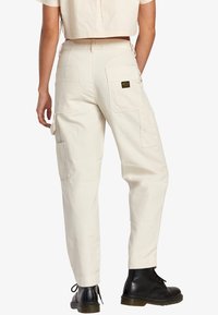 Beige cargobroek met een relaxte pasvorm, voorzien van zijzakken en een achterzak. Gemaakt van katoenen stof. Draagt met zwarte laarzen.