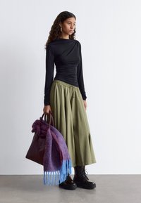 Schwarzes Langarmoberteil, olivgrüner weiter Rock, schwarze Plateaustiefel und eine weinrote Tasche, über die ein blau-lila Schal drapiert ist.