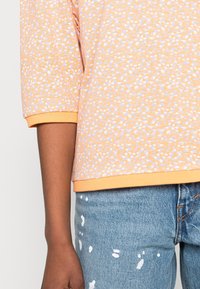 Short-sleeve top in light orange with white and lavender speckles, featuring a ribbed orange hem. Paired with light-wash jeans with white paint splatters.
