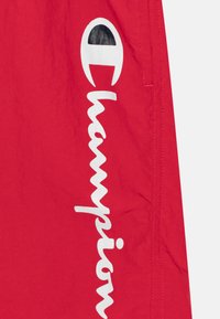 Punaiset shortsit kevyestä kankaasta, jossa on näkyvä valkoinen "Champion"-logo. Sileä pinta ja sivutasku.