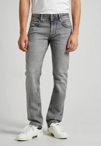 Uomo che indossa jeans aderenti grigi e sneakers bianche, in piedi davanti a uno sfondo semplice.