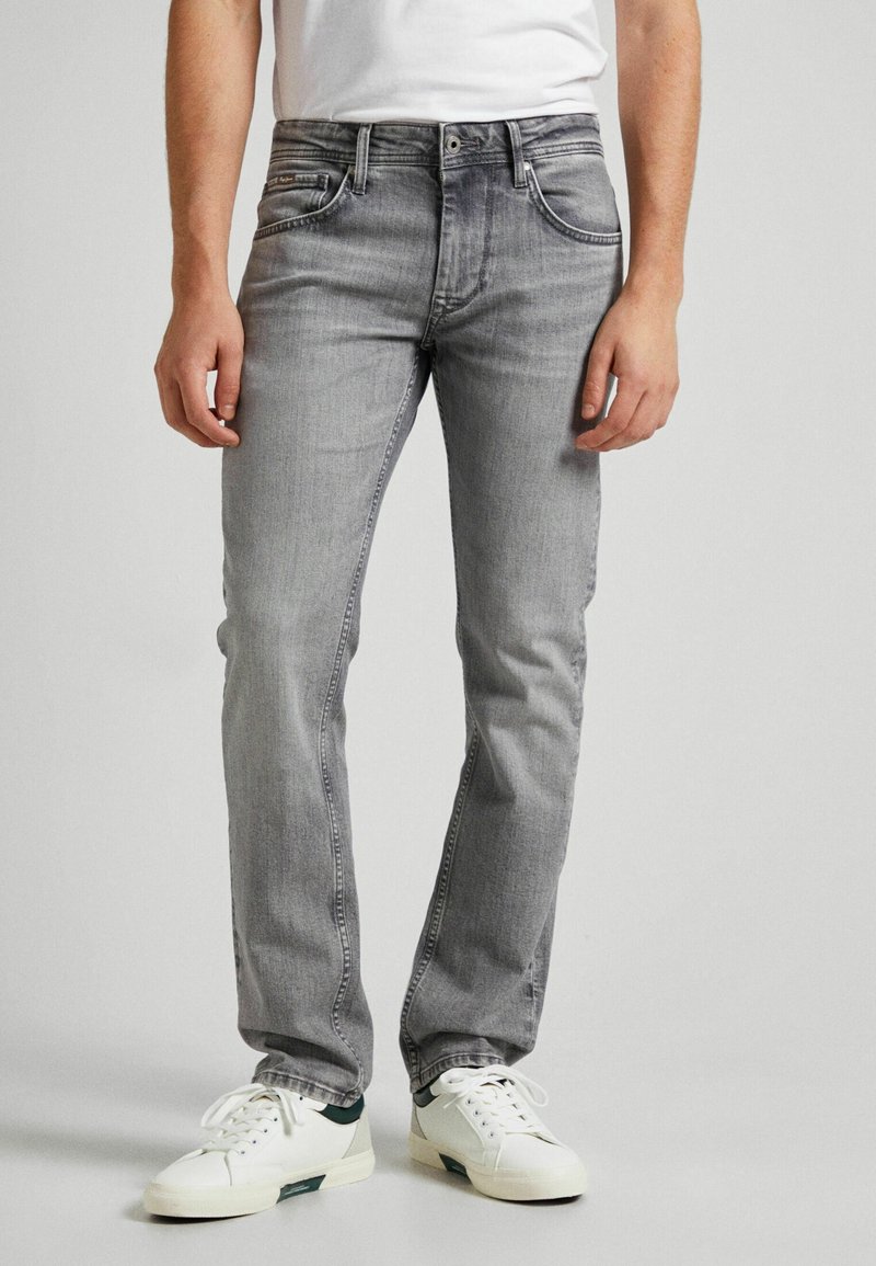 Uomo che indossa jeans aderenti grigi e sneakers bianche, in piedi davanti a uno sfondo semplice.
