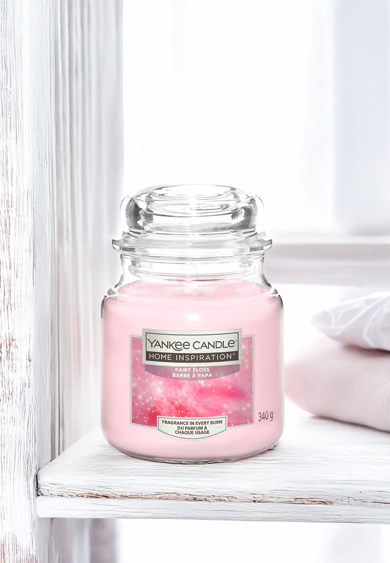 Różowy słoik Yankee Candle z etykietą "Fairy Floss" na białej drewnianej półce obok złożonych białych i różowych ręczników w jasnym pomieszczeniu.