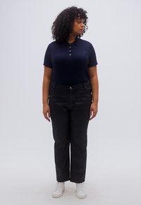 Μπλε ναυτικό πλεκτό πόλο με κοντά μανίκια και τρία κουμπιά, συνδυασμένο με μαύρο τζιν ίσιας γραμμής και λευκά αθλητικά παπούτσια. Απλό φόντο.