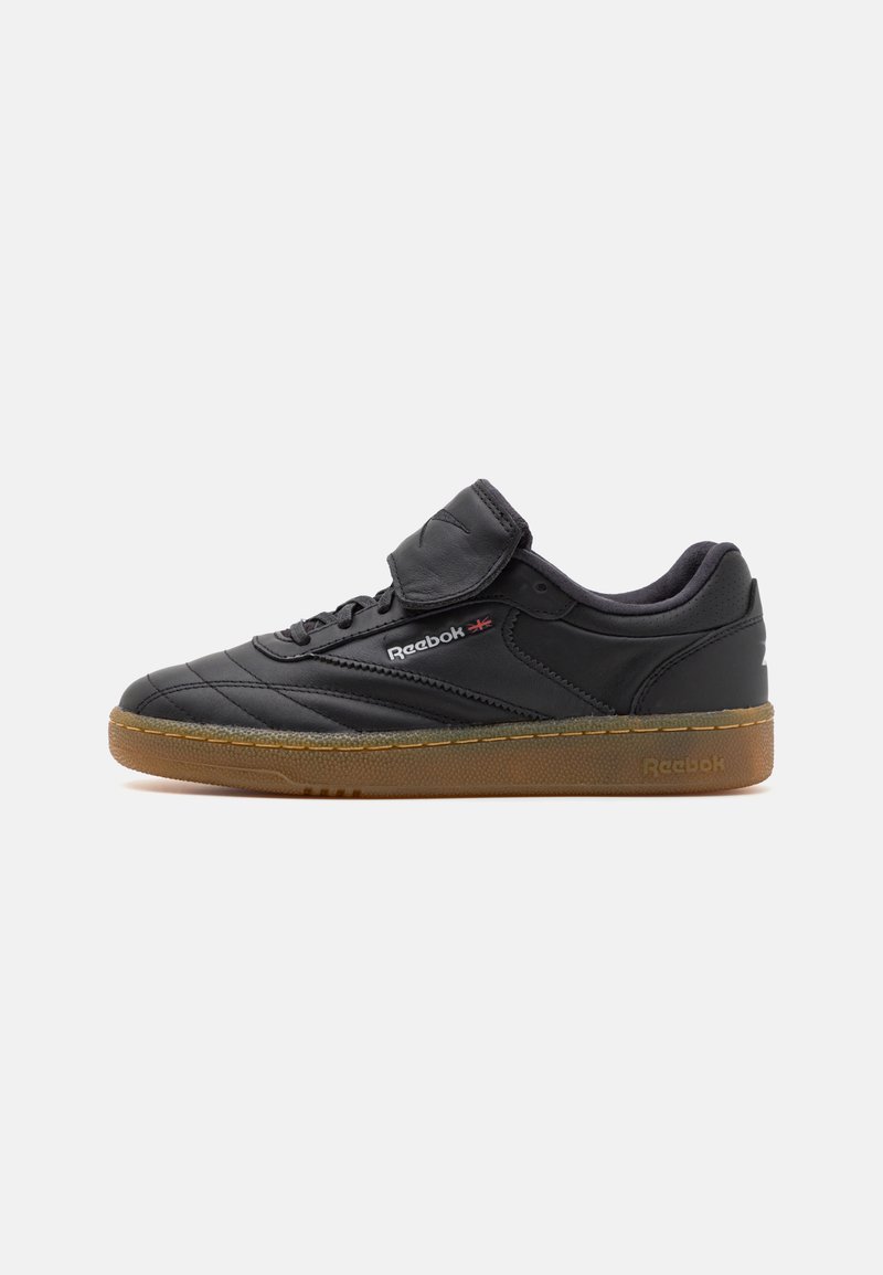 Crne kožne tenisice s podstavljenim jezikom, sa čičak trakama i teksturiranim gumenim đonom. Uključuje Reebok logo sa strane.
