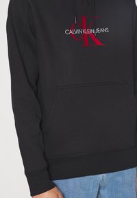 Felpa nera in cotone con tasca frontale, caratterizzata da un audace logo rosso e bianco "CALVIN KLEIN JEANS" e cordini sulla cappuccio.