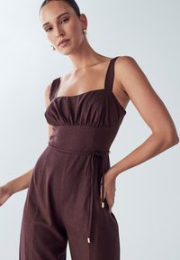 Mono burdeos con un cuerpo ajustado, tirantes anchos, detalles fruncidos y un lazo en la cintura. Tela suave con un acabado ligeramente texturizado.