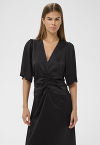 Musthave mustrõhutu must kleit, millel on sügava V-lõikega kaelus, lühikesed puhv-särgid ja kogutud vöökoht. Sile tekstuur ja voolav siluett.