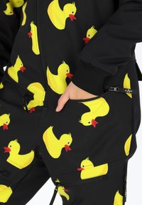 Zwarte jumpsuit met een geel rubberen eendjespatroon, met ritsen op de zakken en ribgebreide boorden. Gemaakt van rekbare stof met een gladde textuur.