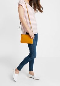 Žlutá kožená crossbody kabelka se zipem, černými kovovými doplňky a nastavitelním popruhem; nošena s pruhovaným topem a těsnými džíny.