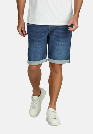 Mann, der dunkelblaue, hochgekrempelte Jeansshorts, weiße Turnschuhe und ein weißes T-Shirt trägt, geht auf einem einfarbigen Hintergrund nach vorne.