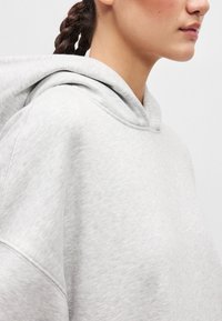 Svetlo siva kapuca s udobnim krojem, mekom teksturom, velikom kapuljačom i suptilnim detaljima šivanja, posebno u području ramena i vrata.