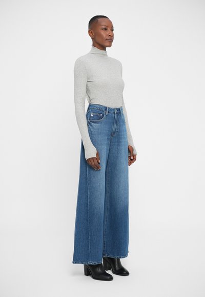 Jeans od plavog denima s visokim strukom i širokim nogavica, nosi se s dugim svijetlosivim puloverom na rolku i crnim čizmama s potpeticom do gležnja.