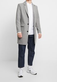 Cappotto lungo grigio con rever a punta, chiusura con un solo bottone e due tasche frontali, indossato sopra una camicia bianca con motivo e pantaloni in denim scuri.