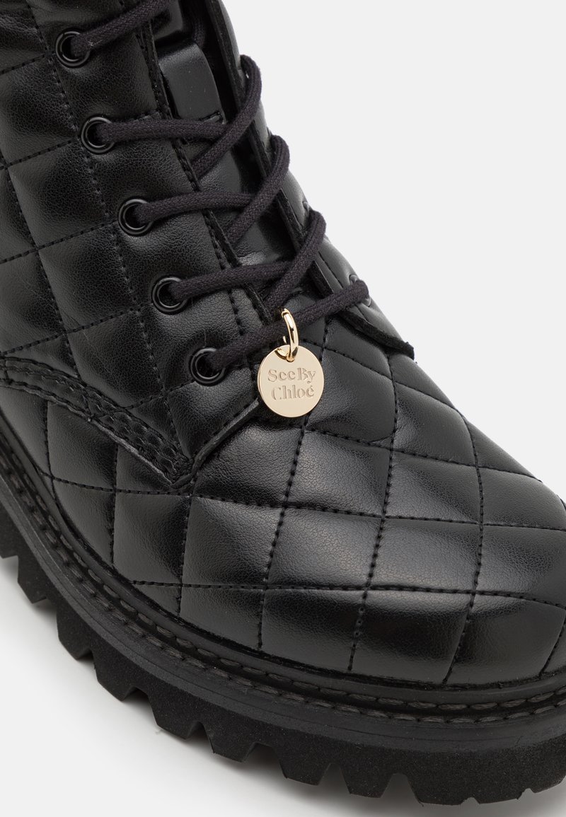 Stivale alla caviglia in pelle trapuntata nera con lacci neri, caratterizzato da una finitura testurizzata e un'etichetta charm dorata. Suola in gomma resistente con bordi frastagliati.