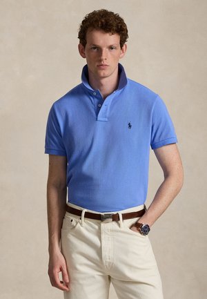 Jonge man draagt een lichtblauw poloshirt, beige broek, bruine riem en polshorloge, staand tegen een effen beige achtergrond.