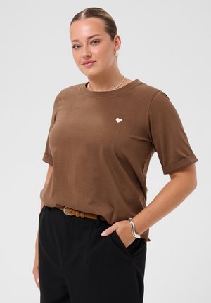Femme avec les cheveux en chignon porte une chemise marron à manches courtes avec un petit détail en forme de cœur, un pantalon noir, un bracelet en argent et une ceinture marron, souriant légèrement.