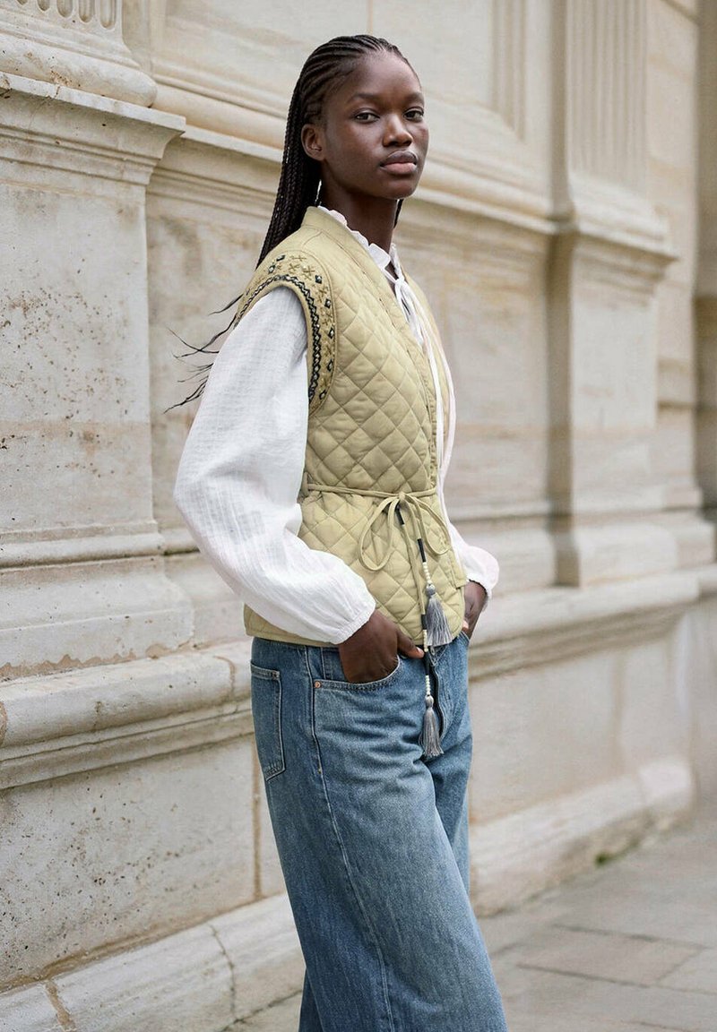 Jeune femme aux cheveux tressés portant un gilet matelassé par-dessus une blouse blanche et un jean bleu, debout contre un mur en pierre, les mains dans les poches.