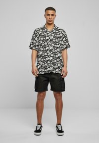 Kortermet, svart blomsterskjorte med hvite blomster; kombinert med svarte cargo-shorts og svarte sneakers med hvite såler.