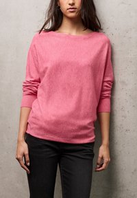 Maglione rosa a maglia testurizzata con scollo ampio e maniche lunghe, abbinato a jeans scuri su uno sfondo neutro in cemento.
