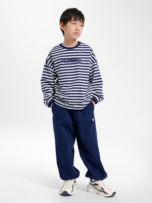Junger Junge in marineblau-weiß gestreiftem Langarmshirt mit "hummel"-Logo, marineblaue Hose, weiße Sneakers, steht mit den Händen in den Taschen.