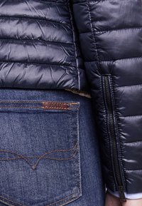 Lähikuva tummansinisestä pehmustetusta takin hihasta, jossa on vetoketju, sinisten farkkujen taskun vieressä, jossa on ruskea nahkainen Polo-tunnus.