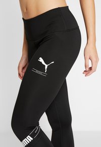 Leggings neri realizzati in materiale elastico, con un logo Puma bianco e accenti testuali. A vita alta, con una finitura liscia e una vestibilità atletica.