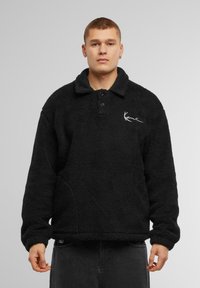 Pullover in fleece nero con colletto, due bottoni e tasche laterali. Presenta un logo bianco discreto sul lato sinistro del petto. Tessuto morbido e testurizzato.