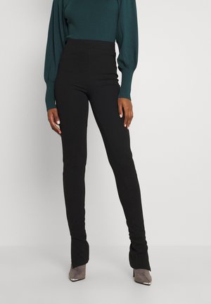 Leggings pretas de cintura alta com uma textura suave, design ajustado e ligeira flare nos tornozelos, combinados com sapatos de salto agudo cinzentos.