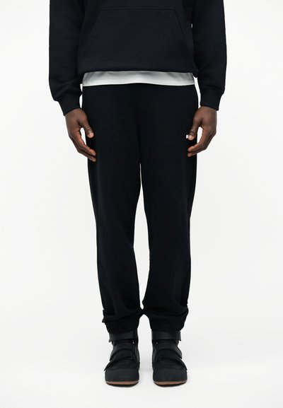 Moitié inférieure d'une personne portant un pantalon de jogging noir, des baskets montantes noires, un sweat à capuche noir et un t-shirt blanc en dessous.