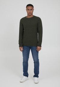 Grønn teksturert strikkegenser med rund hals, kombinert med blå denimjeans og hvite joggesko. Nøytral bakgrunn.