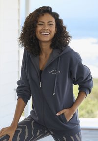 Marineblauwe zip-up hoodie met trekkoorden, voorzakken, geribbelde manchetten en logo-details. Draagt met zwarte leggings met een golvend patroon.