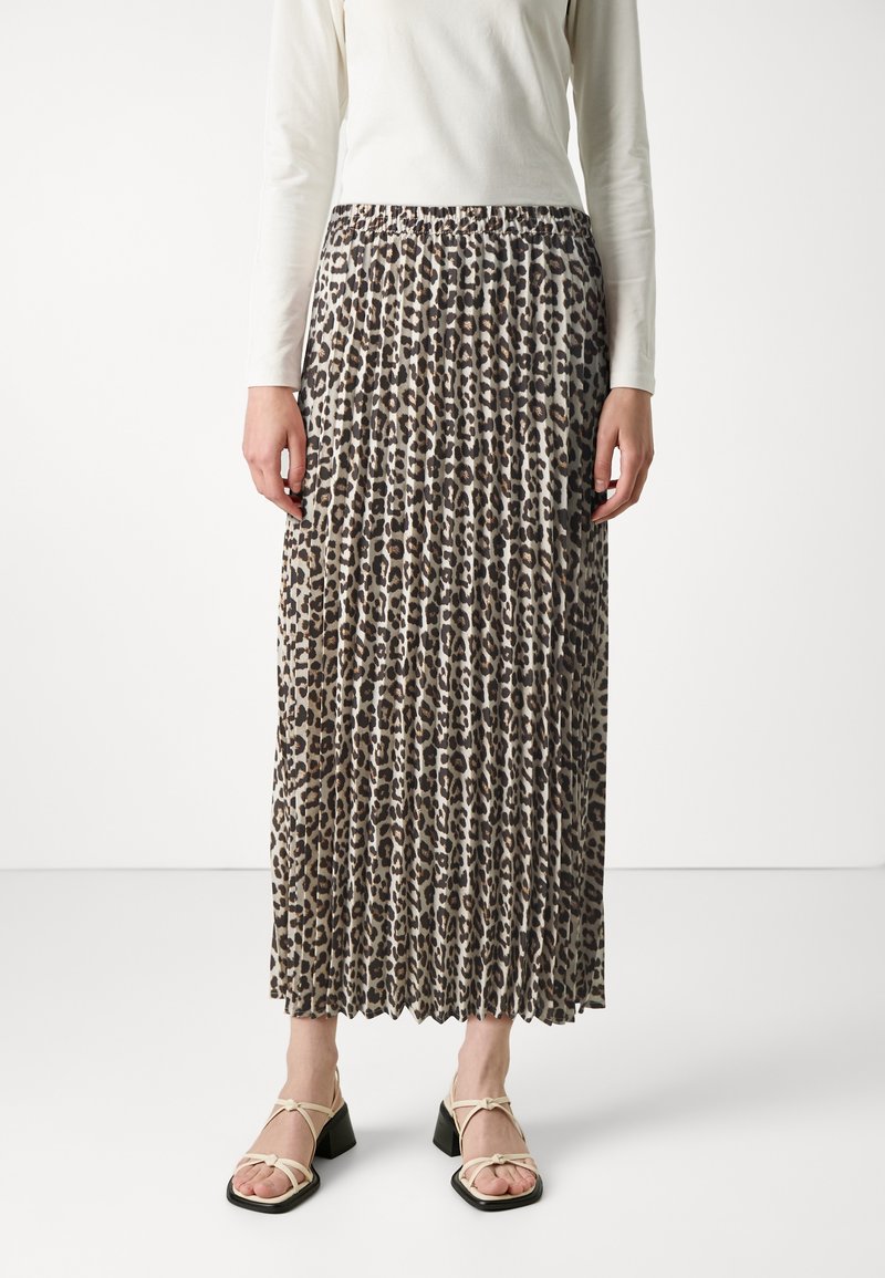 Femme portant une jupe midi plissée imprimé léopard, un haut blanc à manches longues et des sandales à talons ouvertes beige, debout sur un sol blanc.