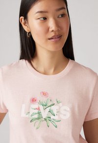 Helles rosa Baumwoll-T-Shirt mit einer botanischen Illustration von Blumen und Blattakzenten, mit dem Text "LEVI'S", der deutlich darunter angezeigt wird.